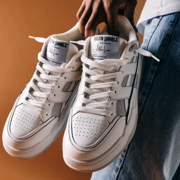 Man houdt witte Austin Gamble Alfa Low sneakers vast voor een oranje achtergrond