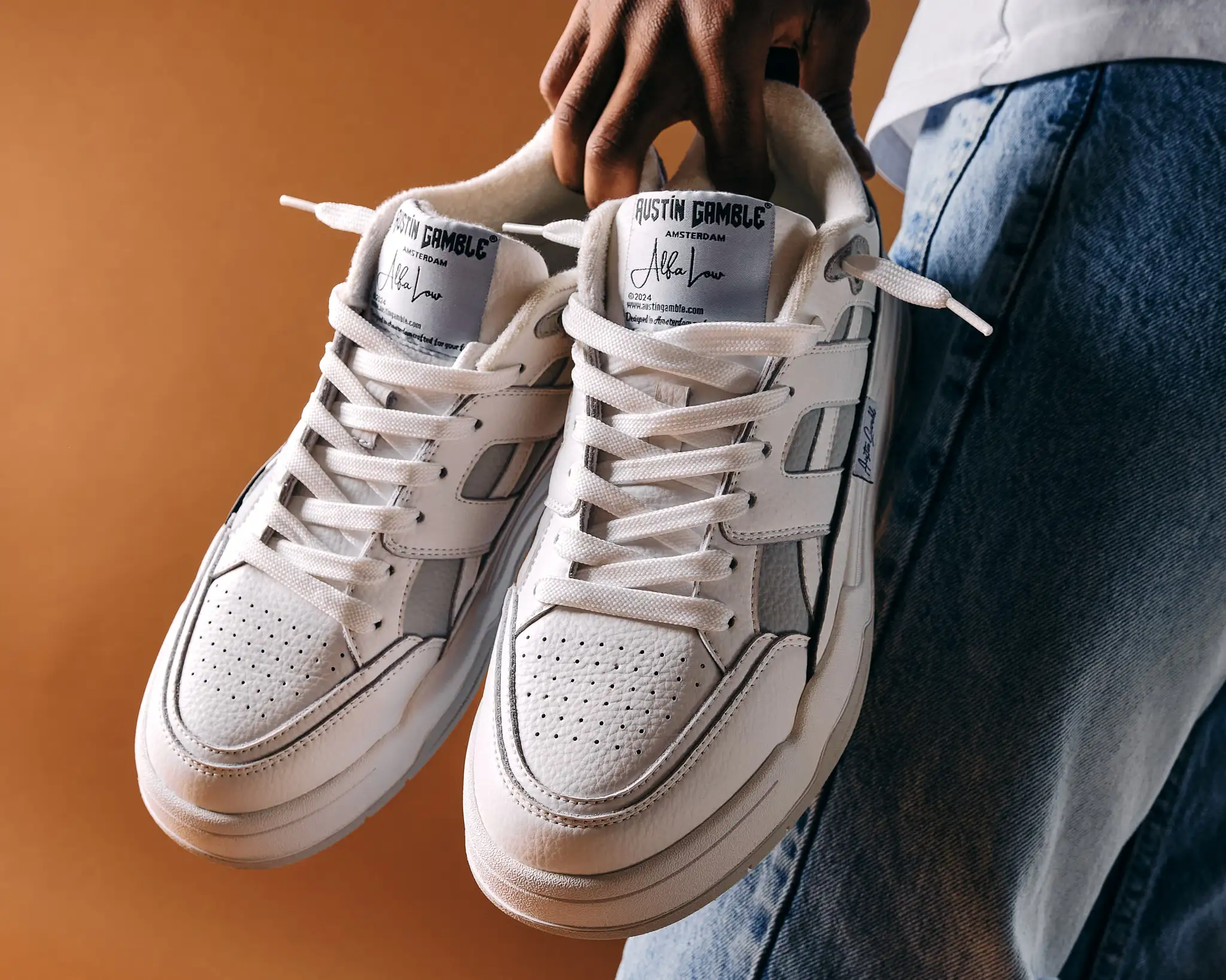 Man houdt witte Austin Gamble Alfa Low sneakers vast voor een oranje achtergrond